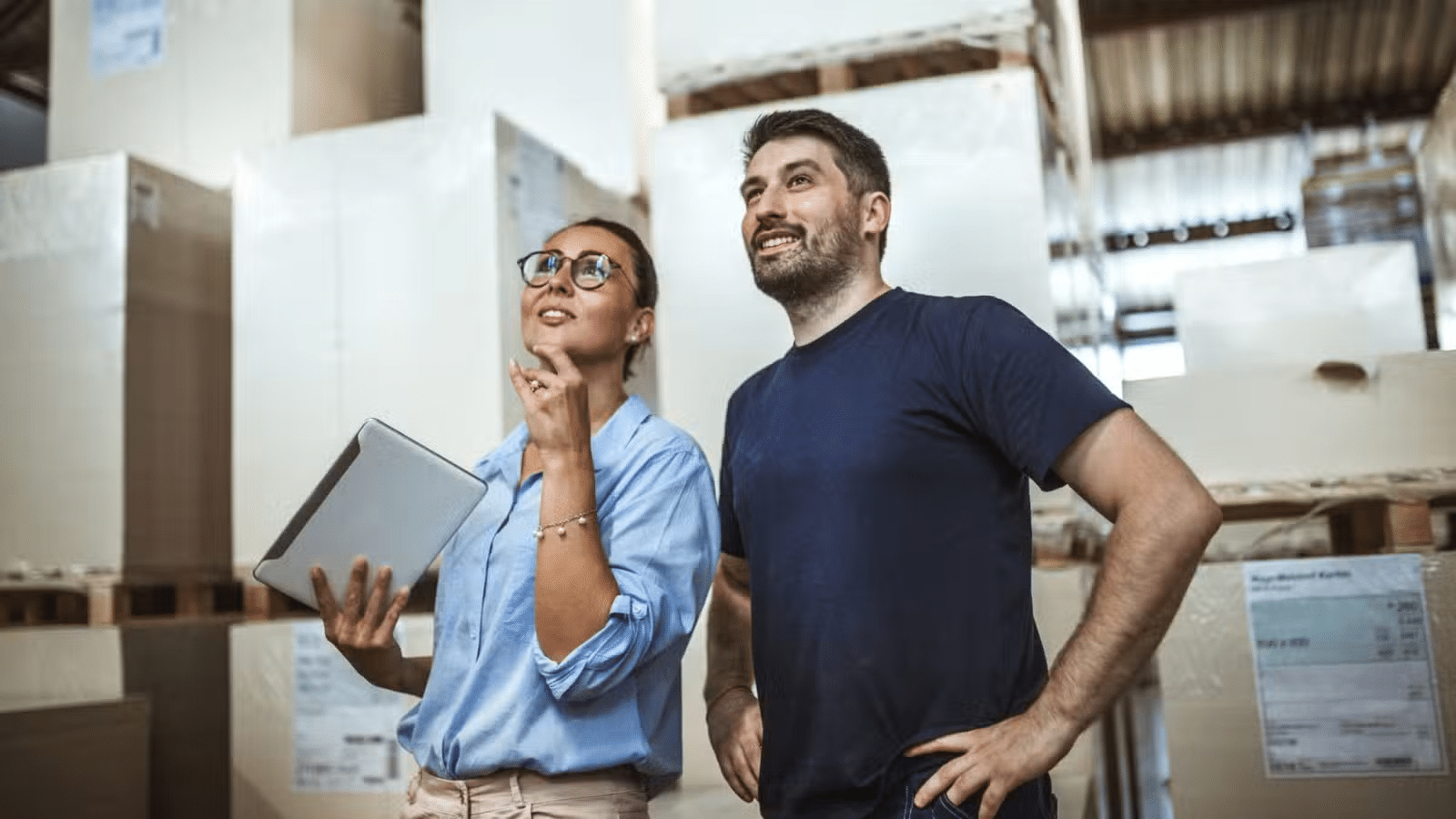 APG-Challenges Two people in a warehouse, looking up and smiling; one holds a tablet, surrounded by large boxes.
