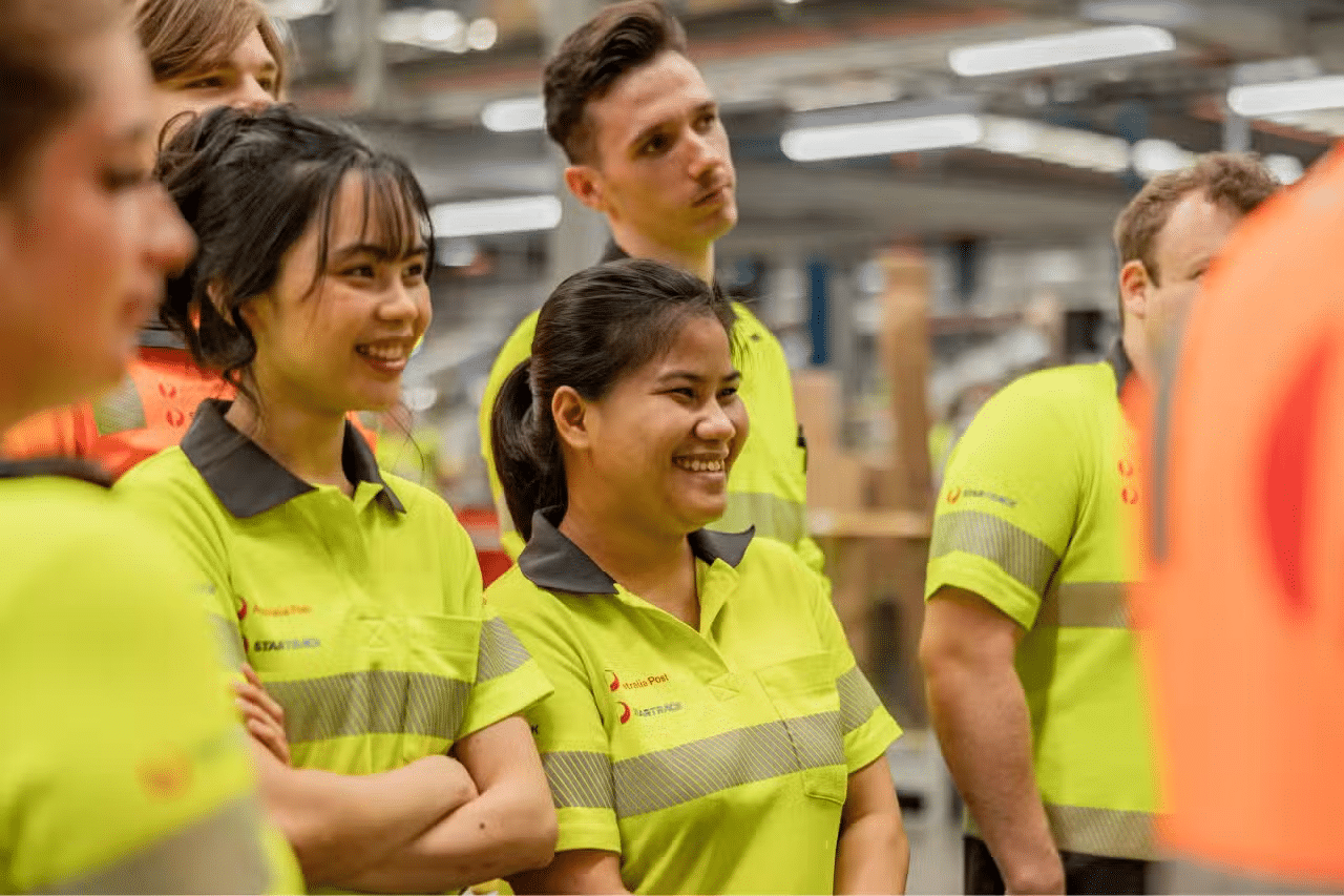 APG-Opportunities Group of people in high-visibility shirts smiling and listening in a warehouse or industrial setting.