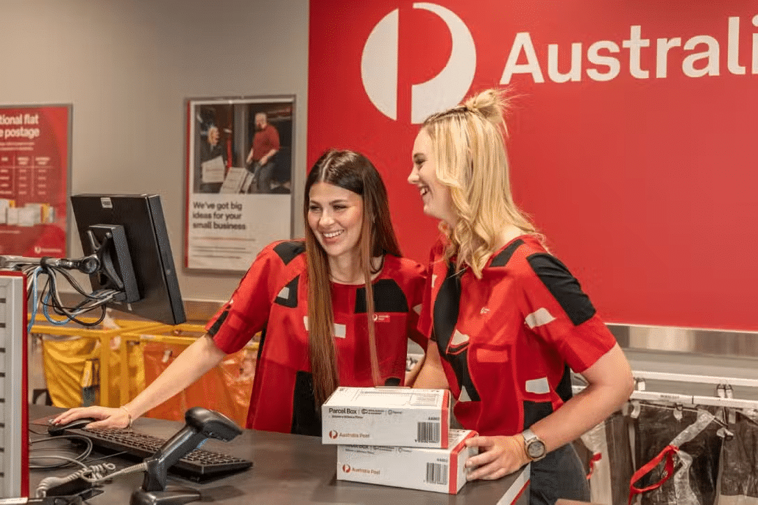 APG-TheImpact Two Australia Post workers smiling behind a counter with parcels and a computer terminal.