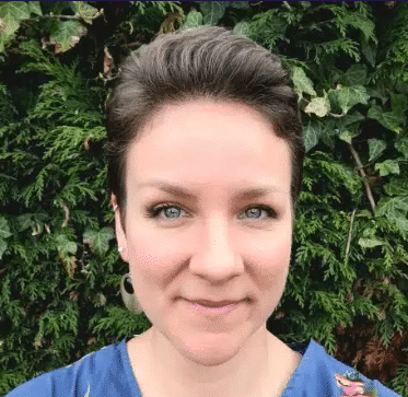 Brit Woman with short brown hair smiling in front of green leafy background.