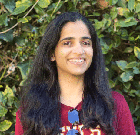 Gouri A woman with long dark hair smiles in front of green foliage, wearing a red shirt and sunglasses.