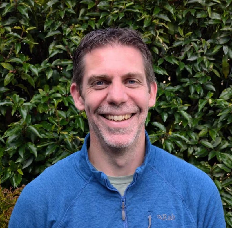 Grant Man smiling outdoors in front of green leafy bushes, wearing a blue zip-up jacket.