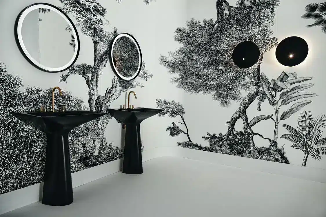 Two black pedestal sinks with round mirrors and a black-and-white forest wallpaper.