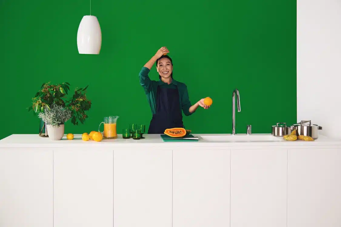 A woman in a kitchen with a green wall, holding an orange and smiling by a white counter and sink.