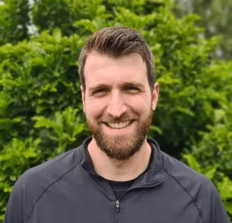 John Man with a beard smiling outdoors in front of green bushes.