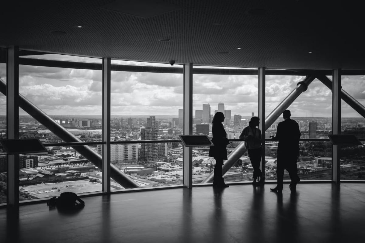 LUPC-Results Three people stand by large windows overlooking a city skyline on a cloudy day.