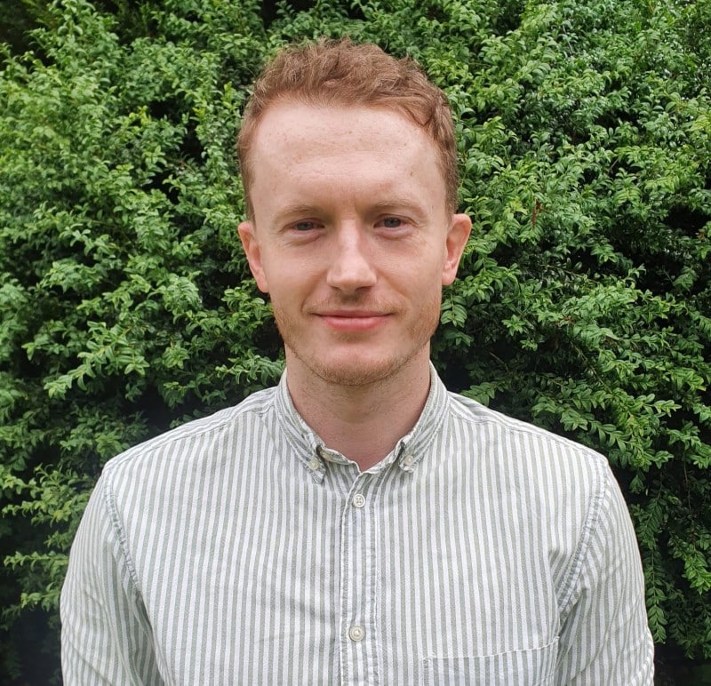 Mike Man with short hair in a striped shirt standing in front of green bushes.