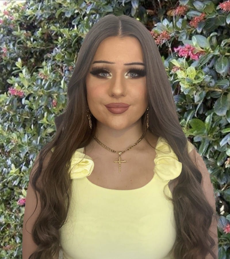 Olivia Woman with long brown hair in a yellow dress stands in front of flowering bushes.