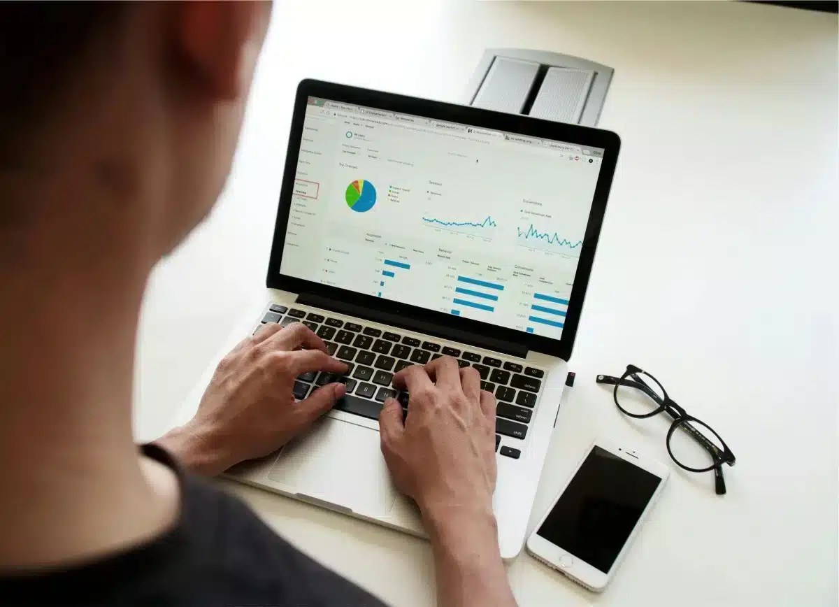 Person working on a laptop with graphs on the screen, glasses, and a smartphone on the desk.