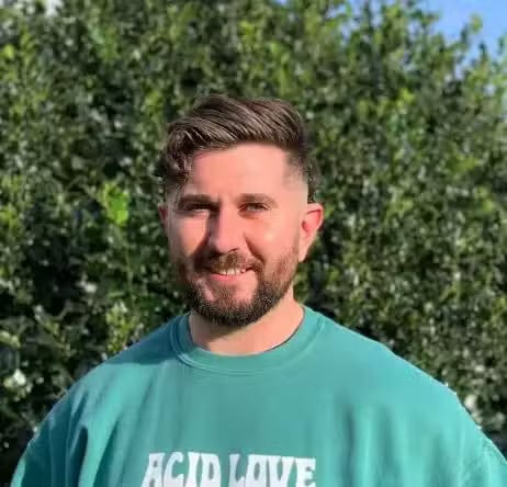 Toby A man with a beard smiles outdoors, wearing a teal sweatshirt, with greenery in the background.