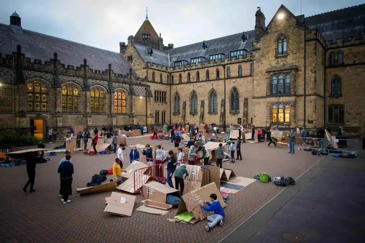Tonbridge-School-Mission Students build cardboard shelters in a school courtyard outside a large historic stone building at dusk.
