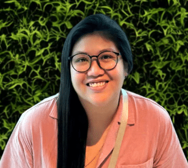 Vina Person with long dark hair and glasses smiling in front of a green leafy background.