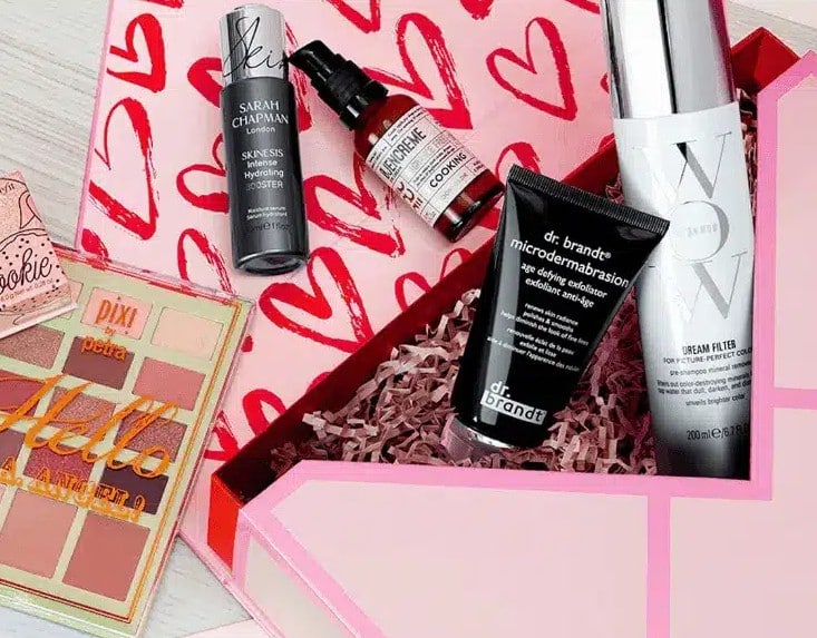 Gift box with various beauty and skincare products arranged on a pink heart-patterned surface.