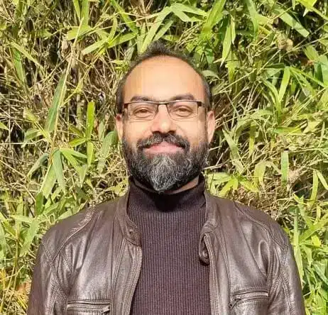 Armughan A man with glasses and a beard stands outside in front of green plants, wearing a brown jacket.