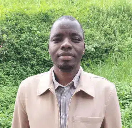 Opio Man standing outdoors in front of green grass, wearing a beige jacket and striped shirt.