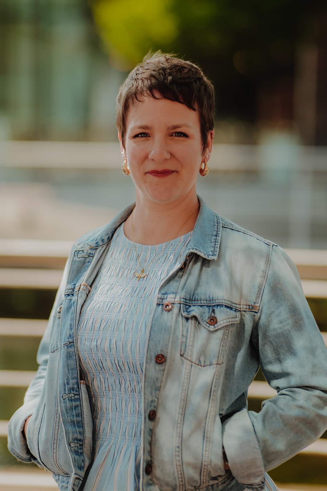 MY CARBON Woman with short hair in a denim jacket, smiling outdoors with a blurred background.