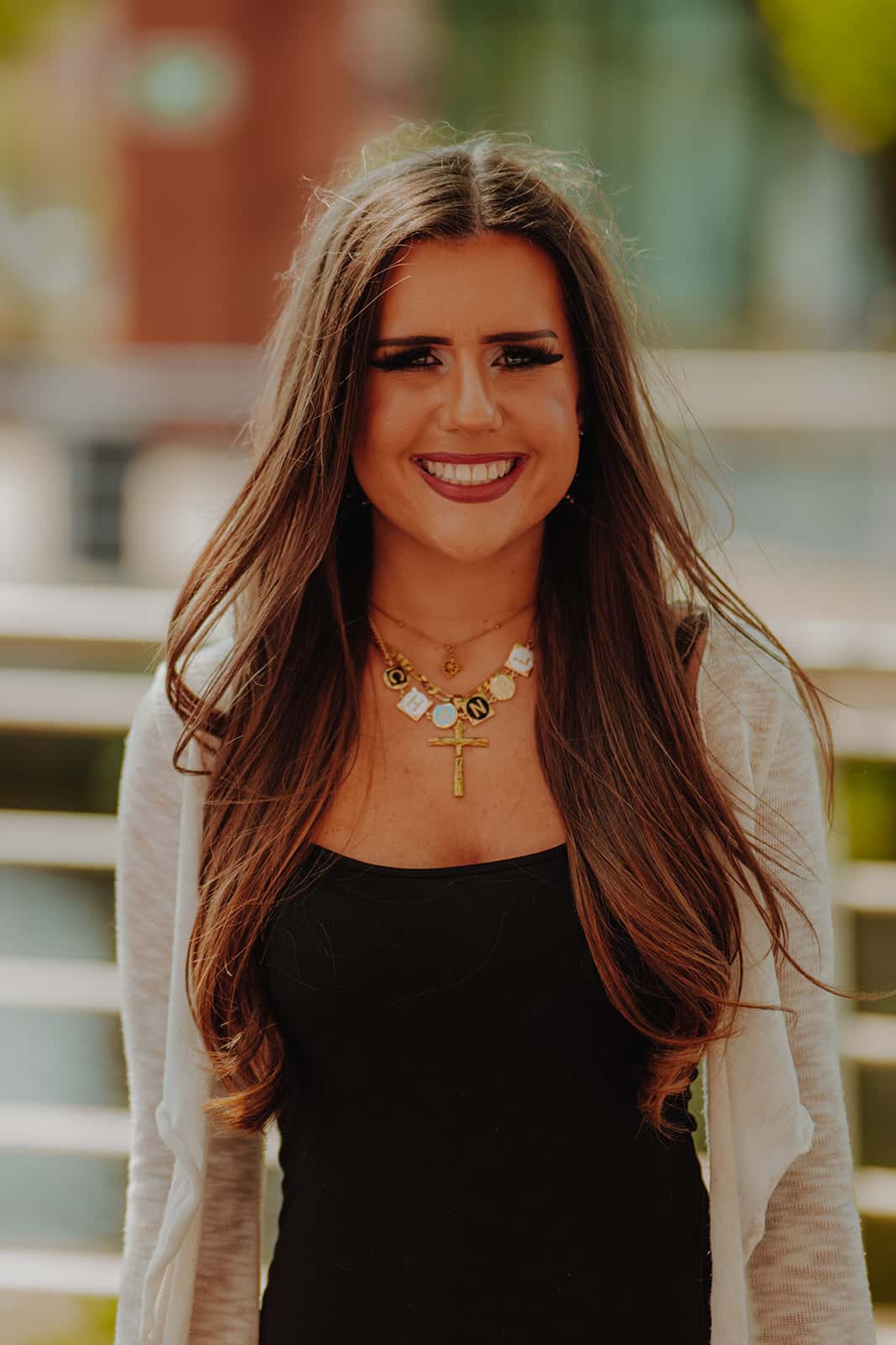 MY CARBON Smiling woman with long brown hair wearing a black top and layered necklaces, outdoors.