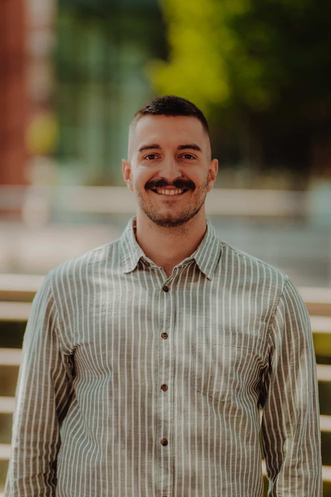 MY CARBON Man with a mustache smiling, wearing a striped shirt, standing outdoors.